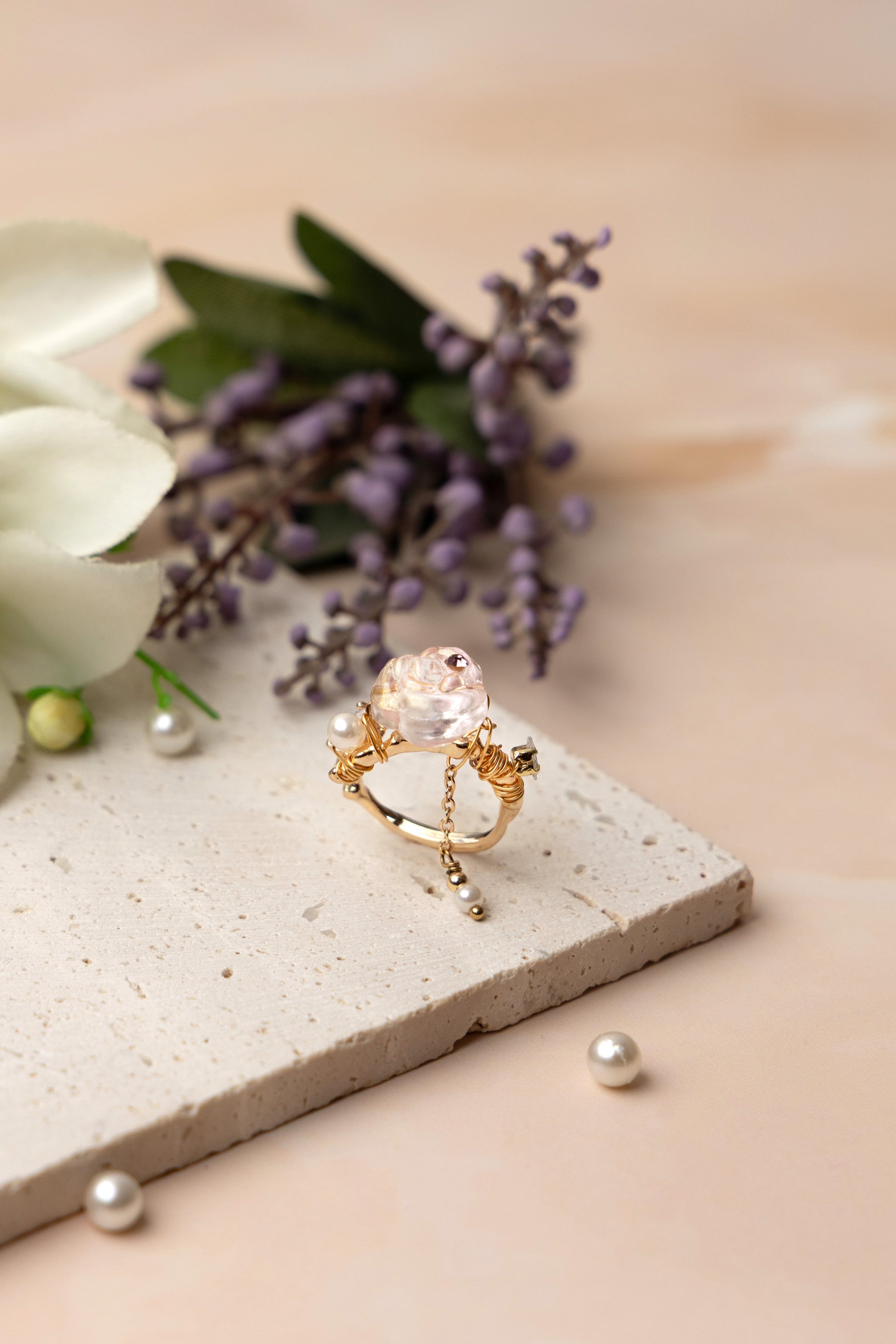 A handcrafted ring with a crystal-like floral centerpiece and an elegant coiled band, featuring a delicate hanging chain with a pearl-like drop that adds graceful movement and distinctive charm