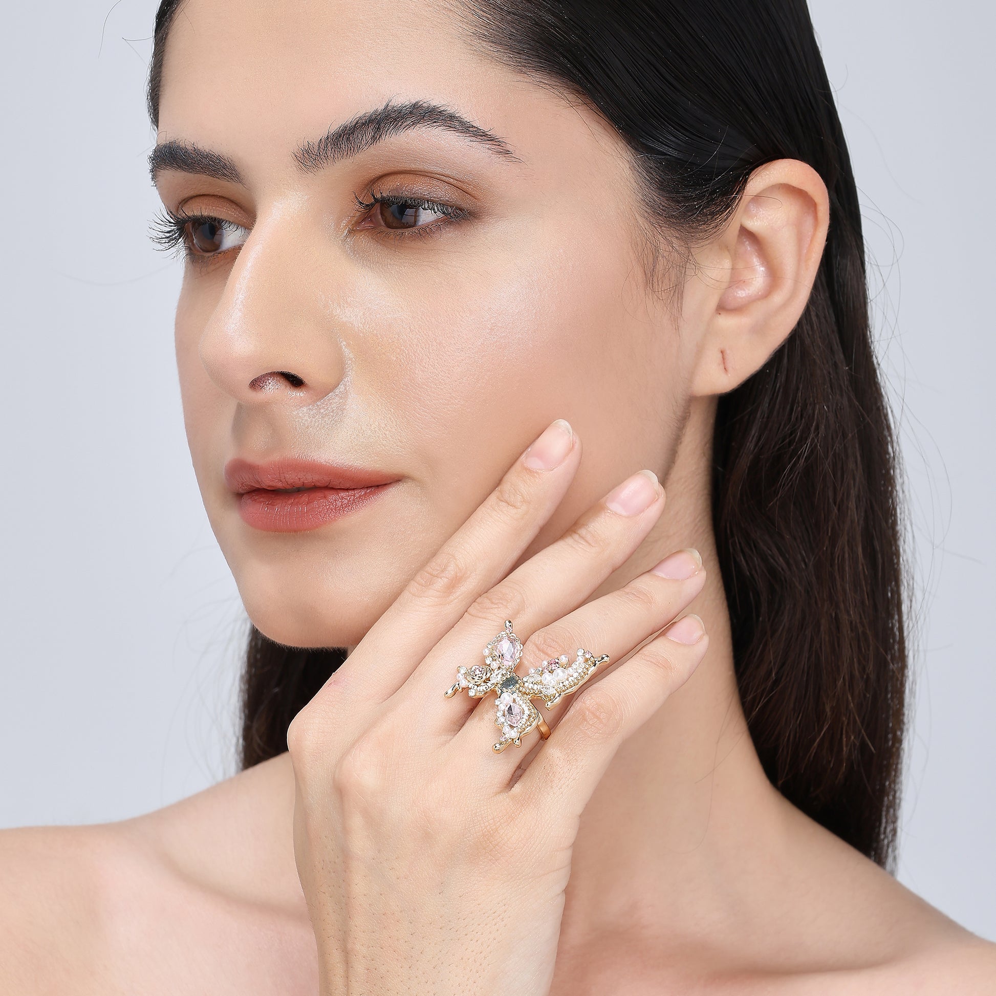 A handcrafted gold-tone ring featuring an ornate butterfly silhouette adorned with hand-placed gemstones, miniature pearls, and crystals in soft rose, mint, and ivory tones, exuding whimsical elegance and sculptural detail.