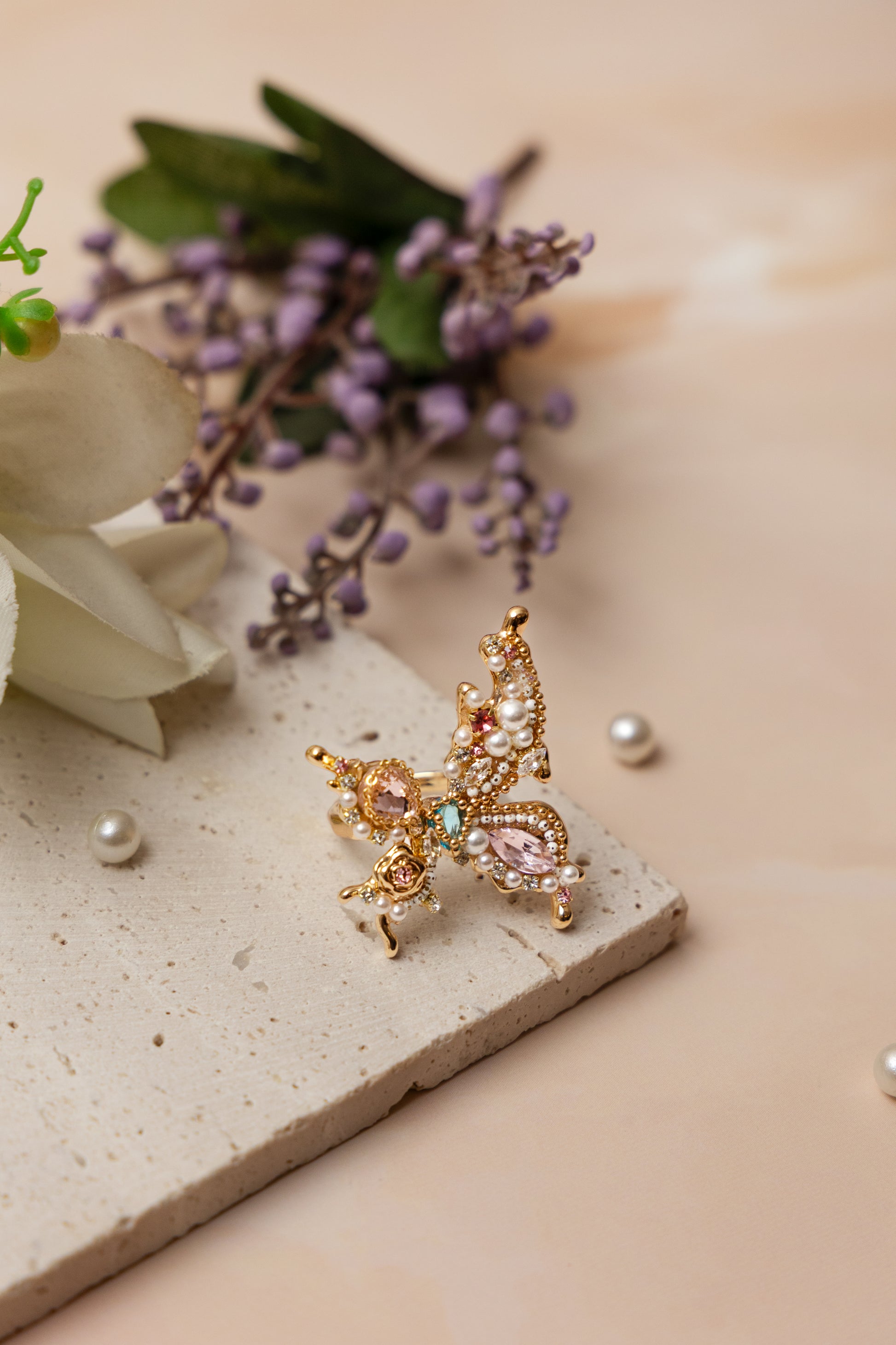 A handcrafted gold-tone ring featuring an ornate butterfly silhouette adorned with hand-placed gemstones, miniature pearls, and crystals in soft rose, mint, and ivory tones, exuding whimsical elegance and sculptural detail.