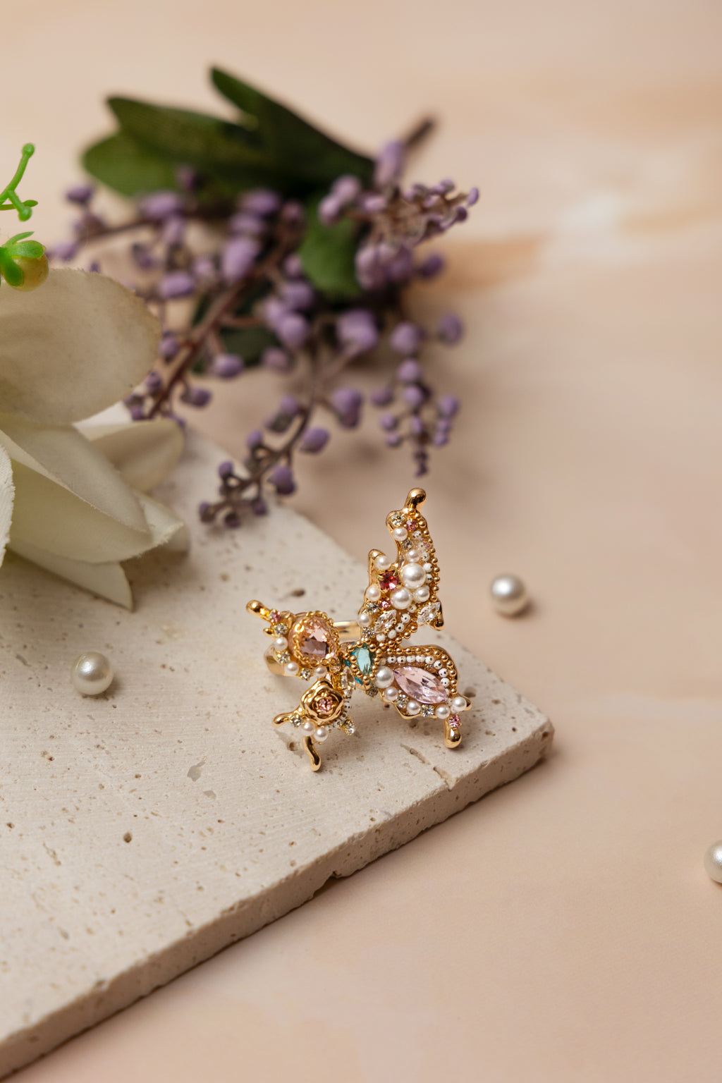 A handcrafted gold-tone ring featuring an ornate butterfly silhouette adorned with hand-placed gemstones, miniature pearls, and crystals in soft rose, mint, and ivory tones, exuding whimsical elegance and sculptural detail.